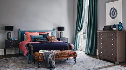 A bedroom with grey walls, teal blue curtains and grey rugs on a wooden floor, colourful red and blue bedding and brown chest of drawers.