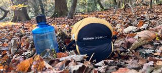 A Forclaz Trekking Sleeping Bag MT900 5°C Down in its stuff sack on a leafy woodland floor, next to a large water bottle.