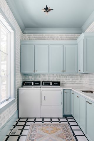 pastel blue laundry room with painted ceiling, cabinetry, wallpaper, black and white tiled floor, rug