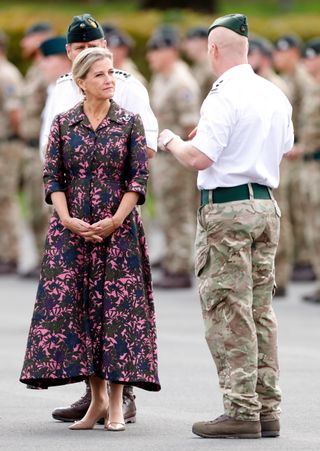 Duchess Sophie wearing a long pink and blue patterned dress and clasping her hands in front of her while talking to two soldiers outside