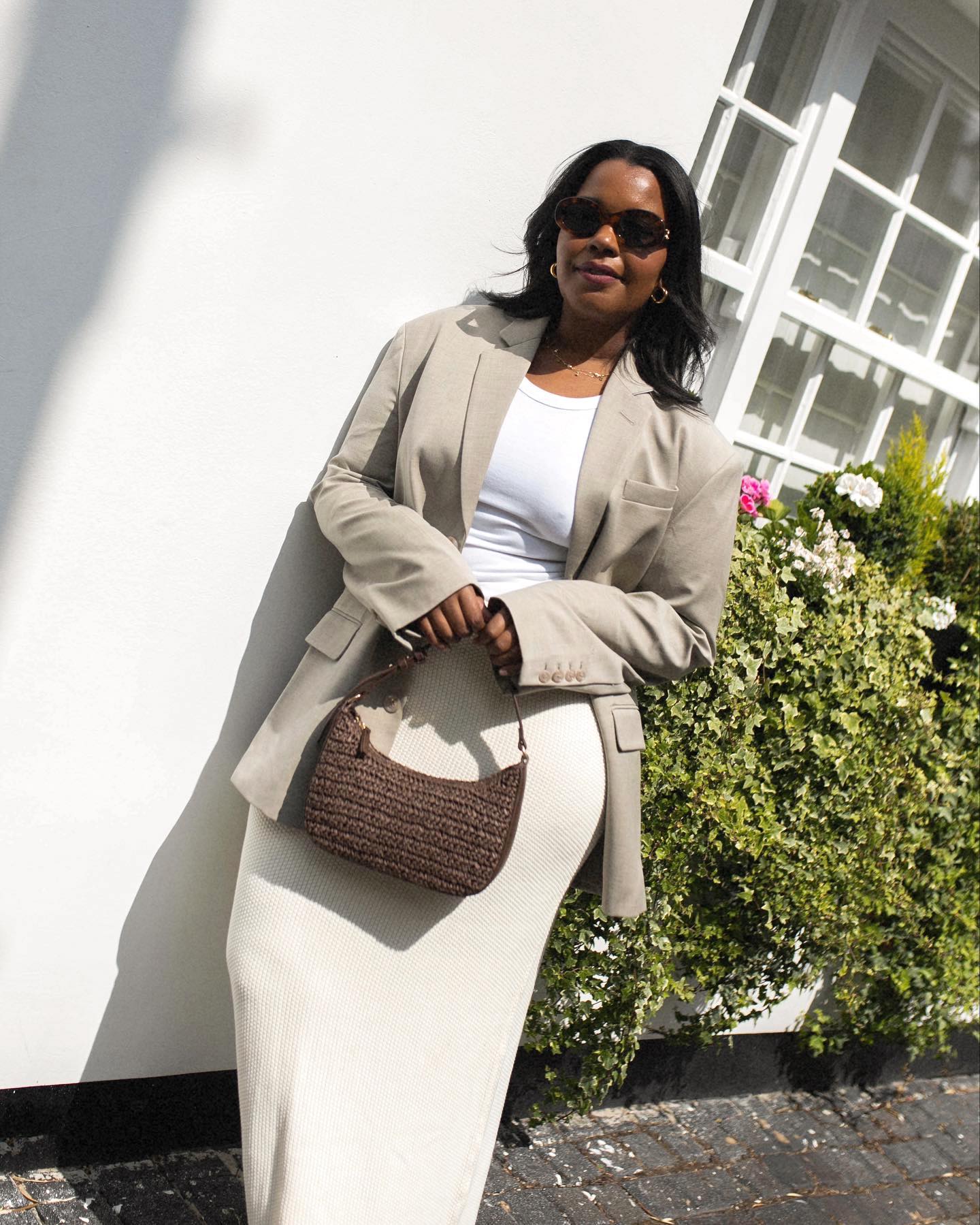 Fashion influencer Karina of Style Idealist leans against a white wall next to a garden wearing tort oval sunglasses, a neutral tan blazer, white t-shirt, cream maxi skirt, and a woven brown shoulder bag.