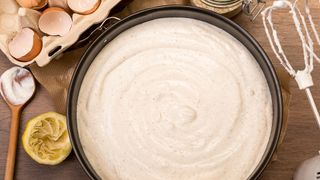 A cake tin filled with cake batter, with used eggs, lemon rind, whisk and wooden spoon beside it