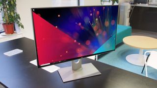 Dell UltraSharp UP3218K på et skrivebord med en plante og møbler i baggrunden