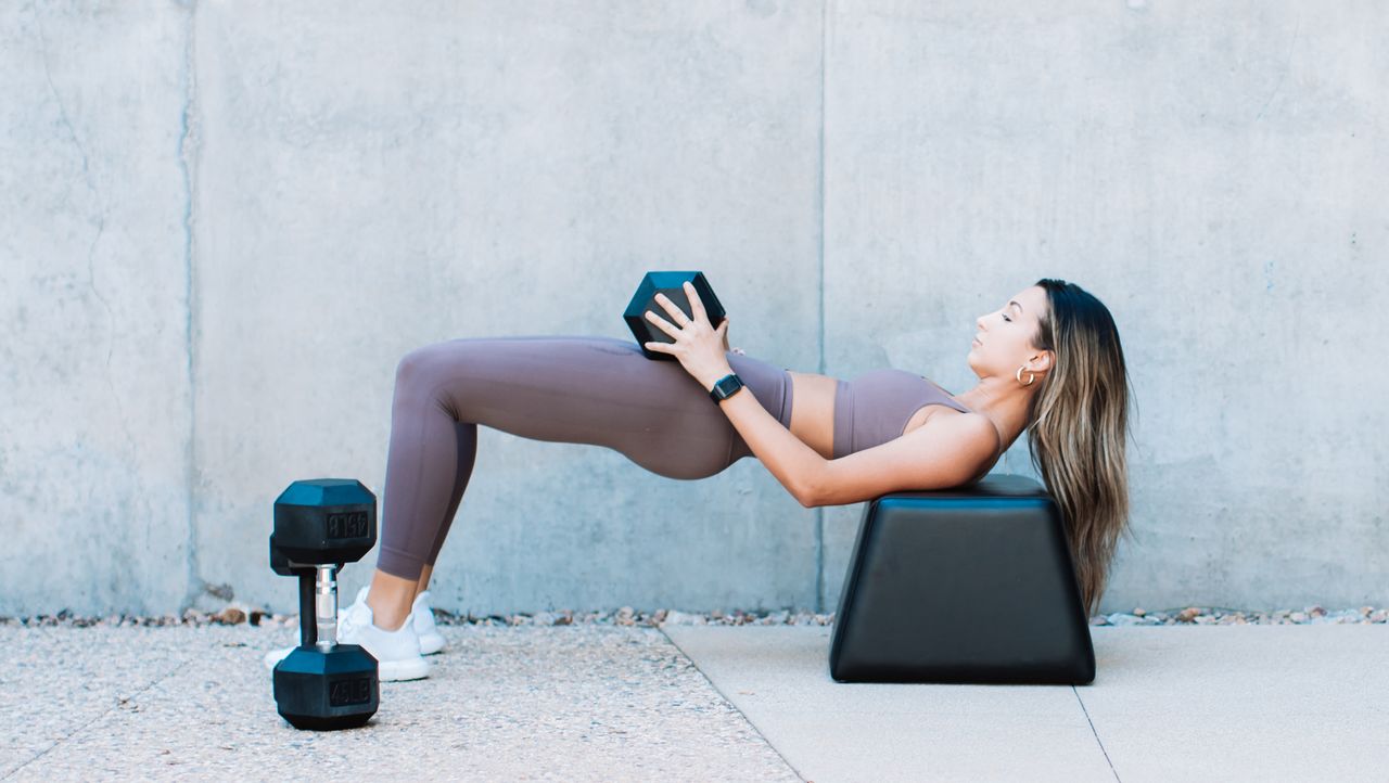 Woman exercising outside next to a grey concrete wall. Her back rests on a bench shaped as a trapazoid, her hips are raised and a dumbbell rests on her hips. Her feet are flat on the floor. 