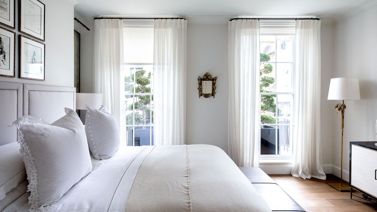 Mattress topper vs. new mattress. Double bed in pale grey bedroom with wooden floor, cream and white bedding and white curtains dressing floor to ceiling windows.