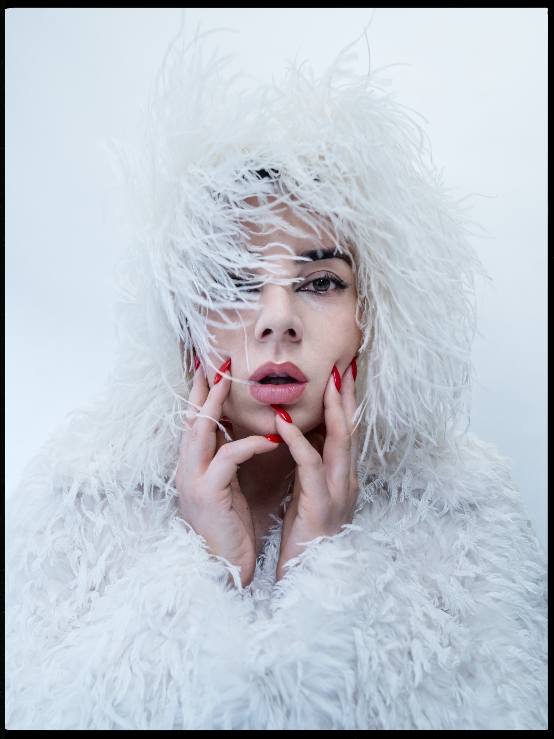 Close-up of Shon Faye wrapped in a white feathered hood and coat, with red pointed nails touching her face and feathers falling across one eye