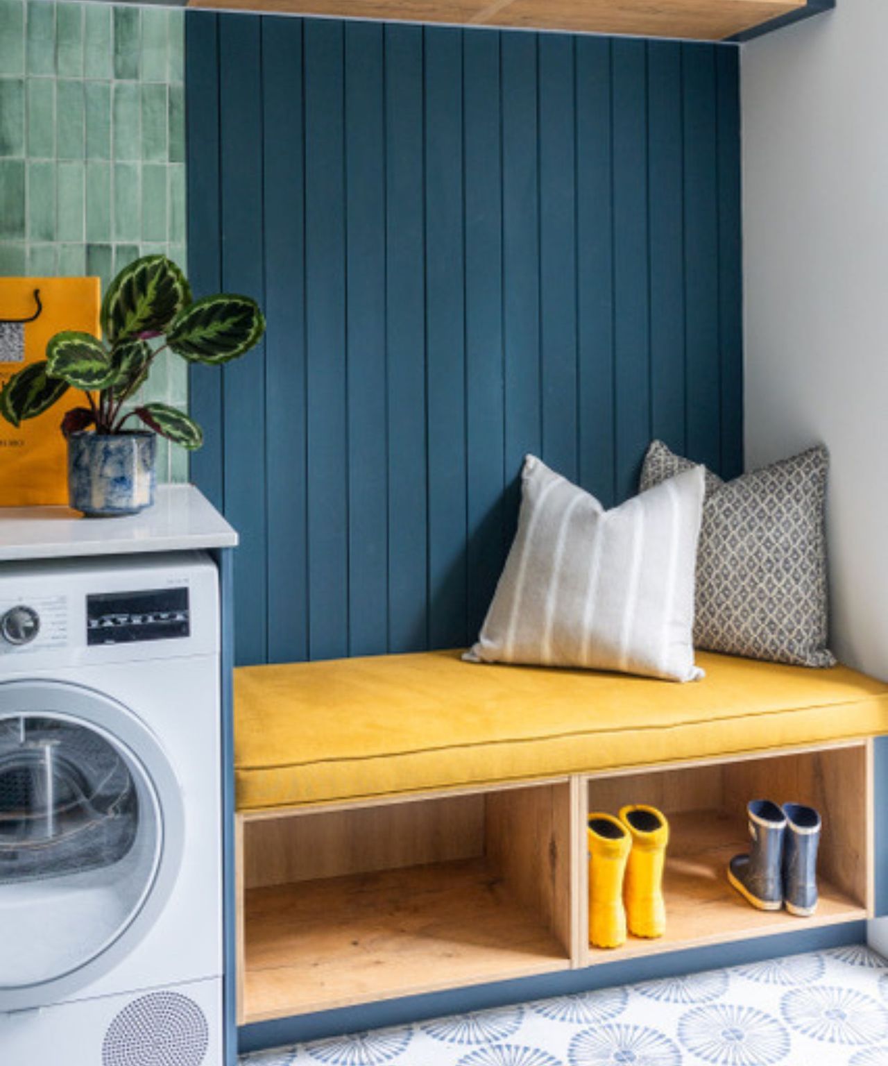 A utility room with green glazed square wall tiles, dark blue tongue-and-groove panelling, a yellow cushioned bench with shoe storage, patterned floor tiles and a washing machine.