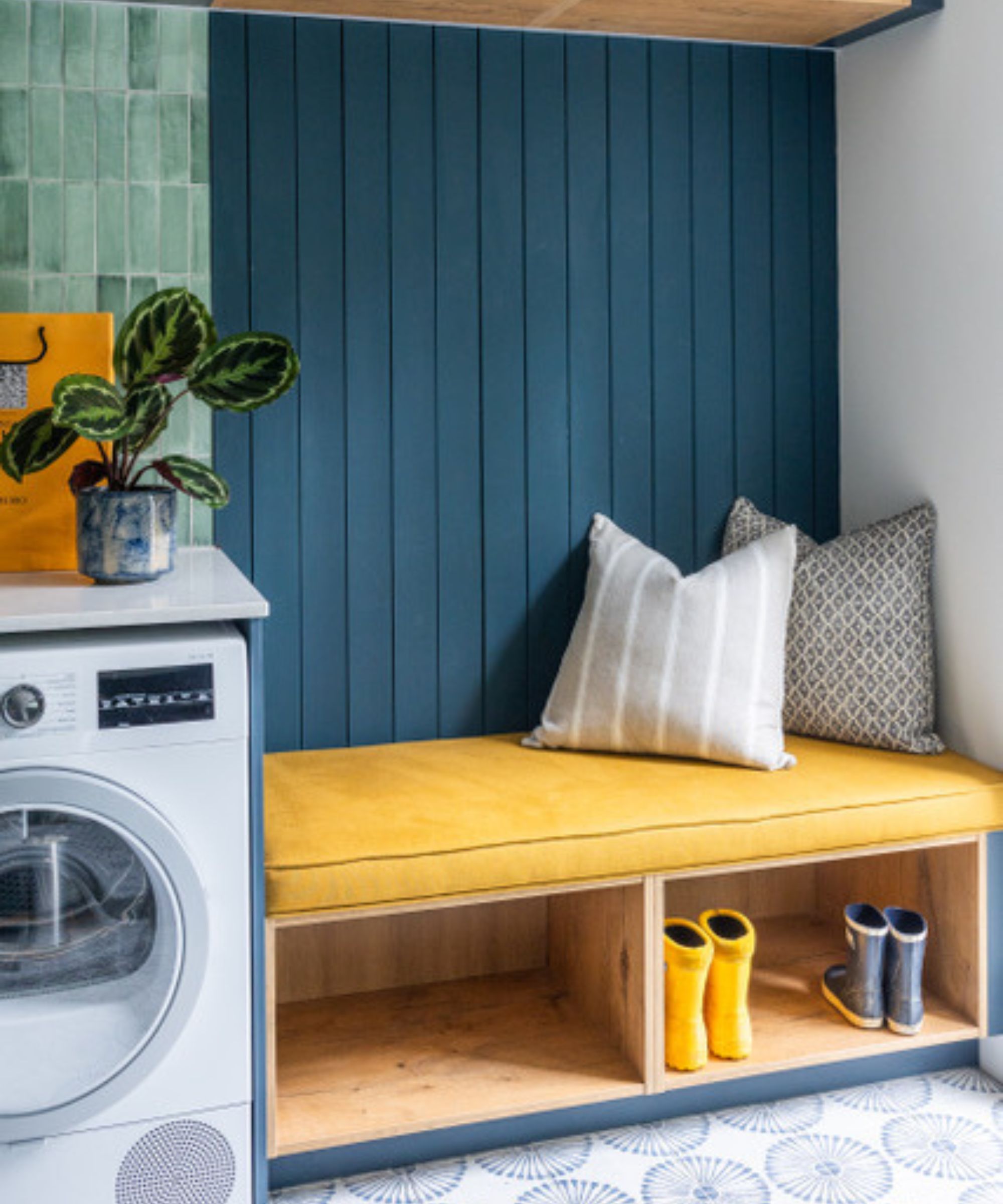 A utility room with green glazed square wall tiles, dark blue tongue-and-groove panelling, a yellow cushioned bench with shoe storage, patterned floor tiles and a washing machine.