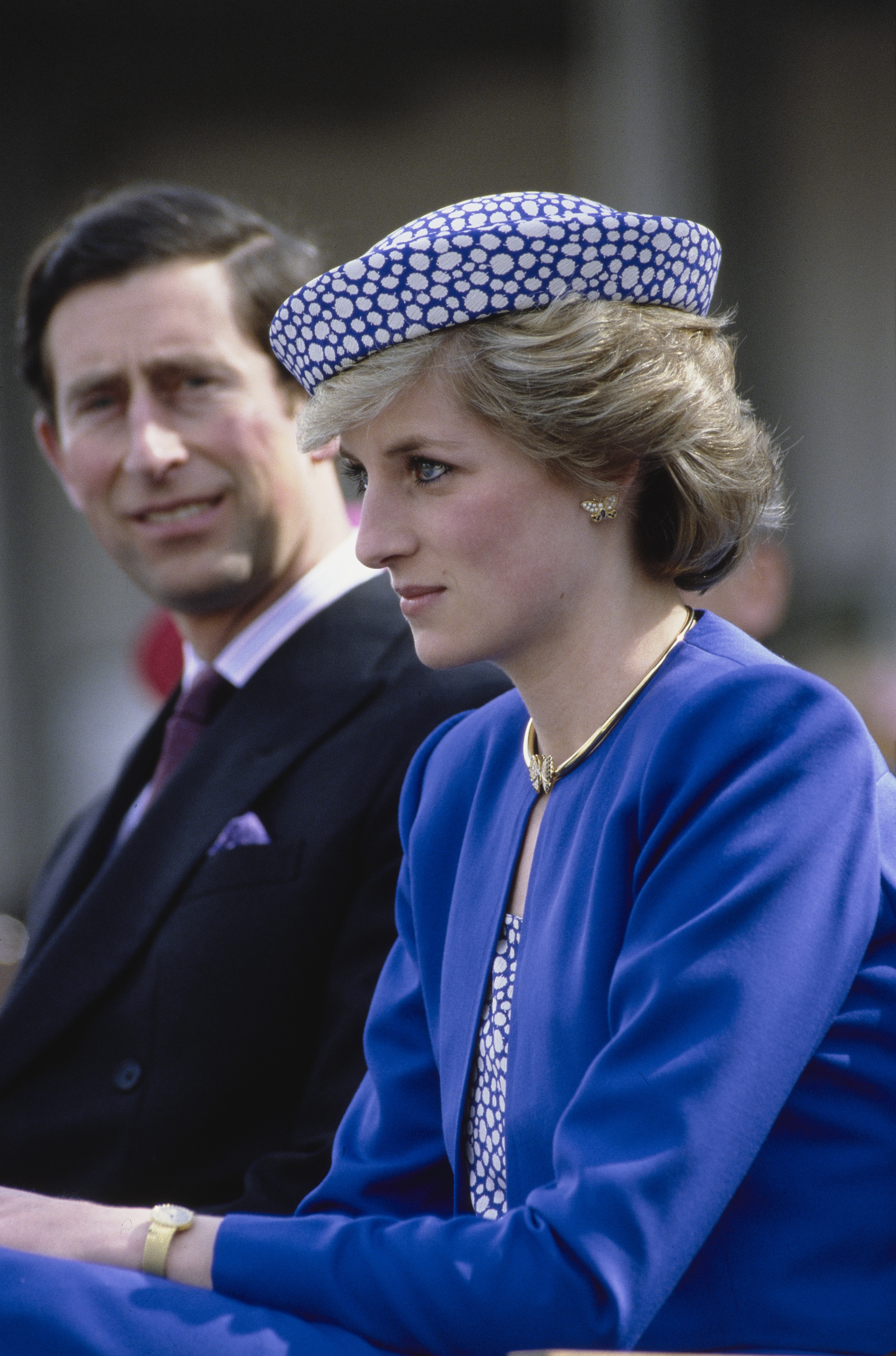Princess Diana wearing a blue jacket and polka dot hat sitting next to Prince Charles