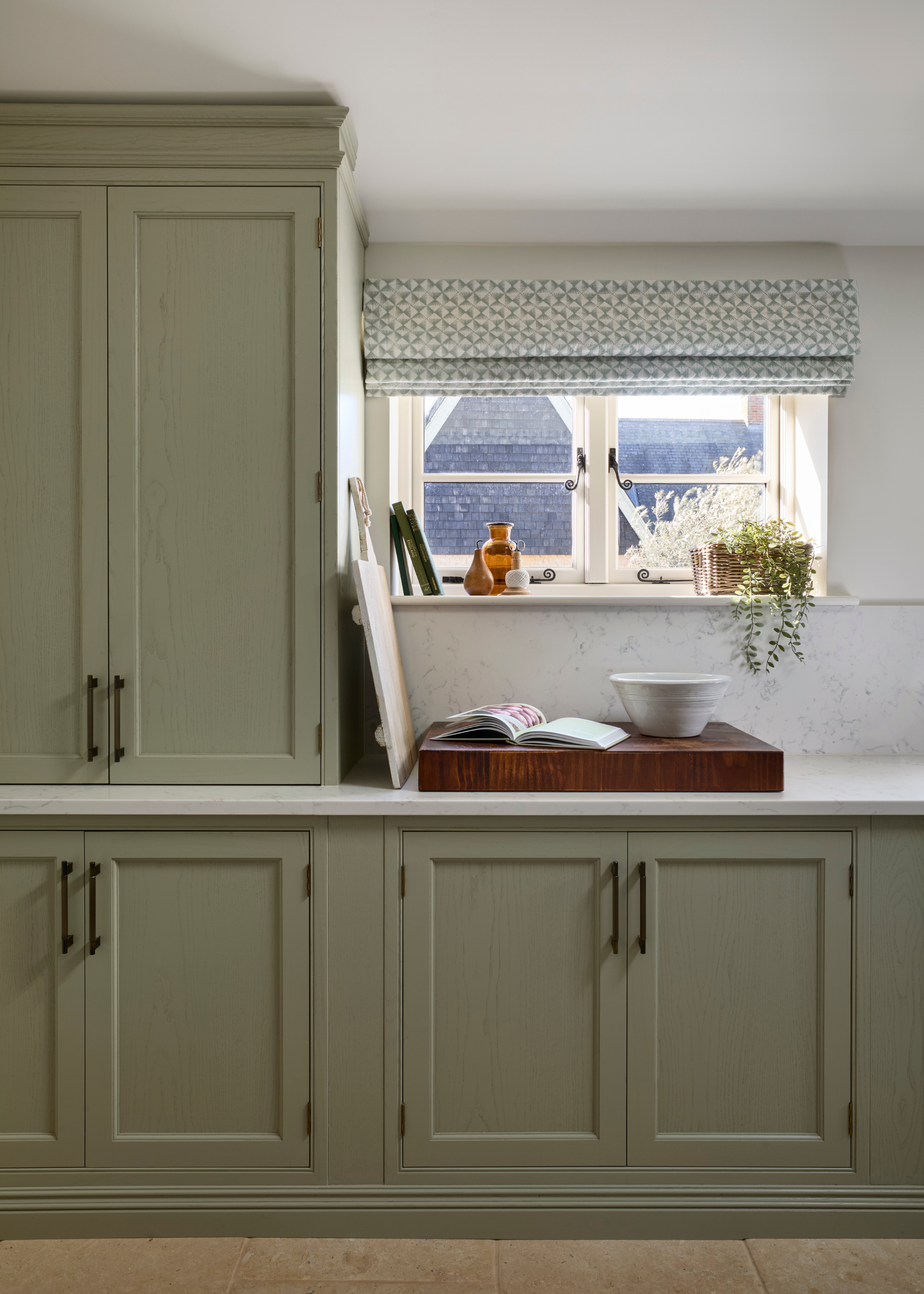 A traditional sage green kitchen with a small window and light blue roman blind hanging over it