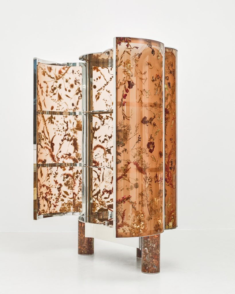 A cabinet featuring a resin body with chrome detailing and encapsulated, frozen-in-time florals, petals, and vegetation inside it, showcased in a brightly lit gallery setting.