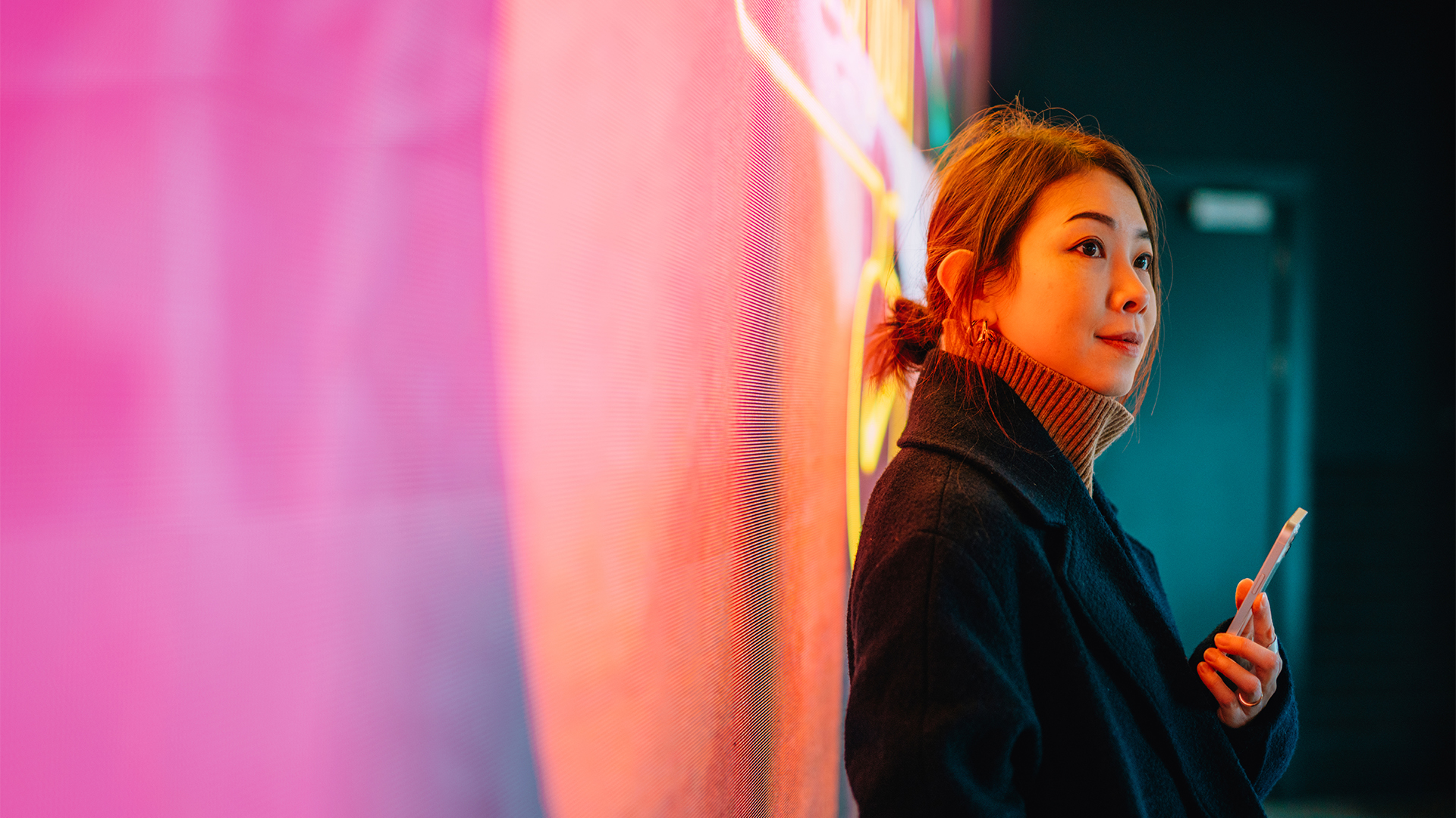 Young Asian woman holding smartphone while leading back against a bright-colored orange and purple wall.