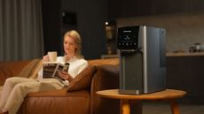 The Waterdrop A1 filtration system on a side table beside a leather sofa, where a woman sits drinking a coffee.