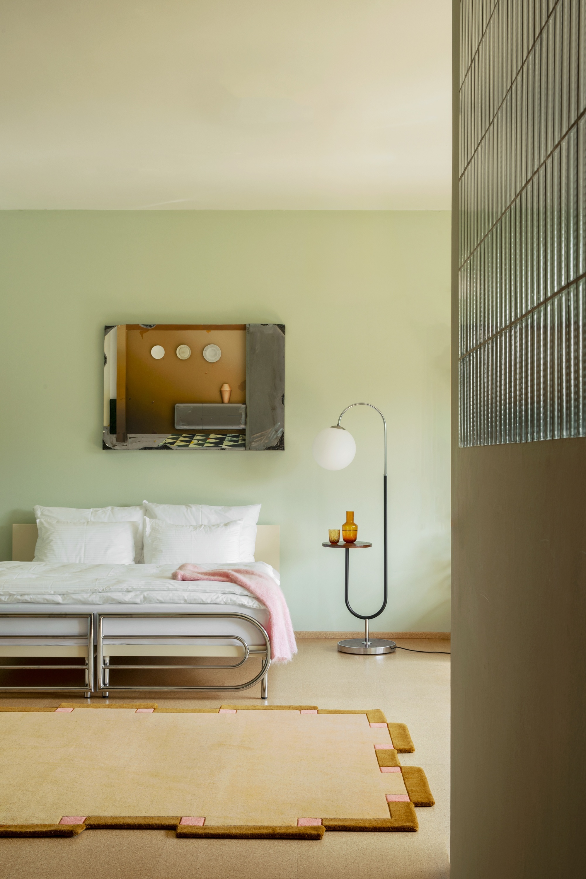 Image of a mint green bedroom with a modern bed that has a silver frame and white bedding. The floors are tan with a tan rug that has a caramel and pink border.