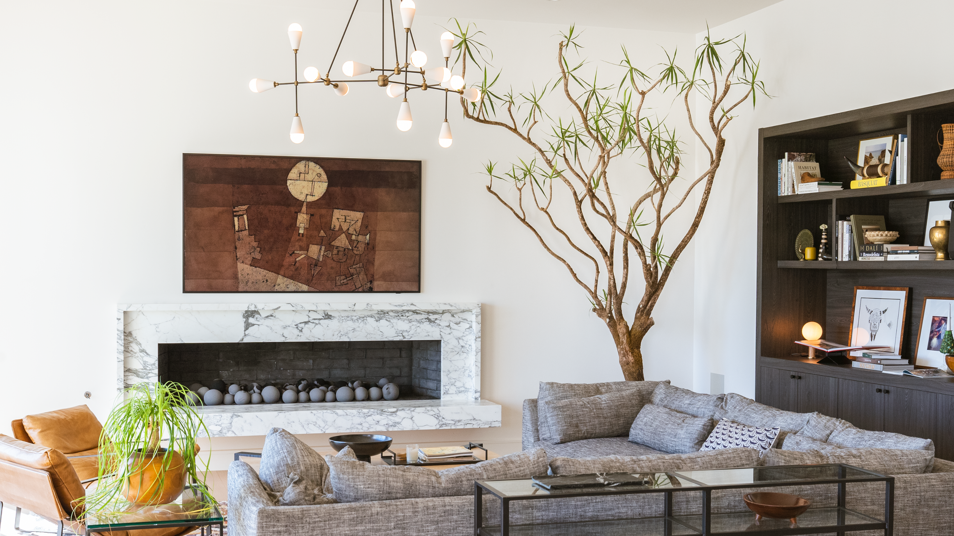 A modern living room with a large sculptural indoor tree in the corner that commands the space, next to an inset black bookcase on the right and an inset marble fireplace in the center with a gray l-shaped sofa facing it