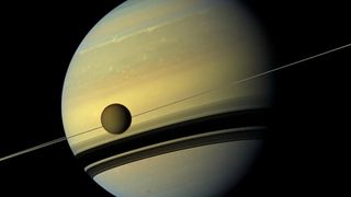 saturn hangs huge, the shadows of its rings striping its lower half. a moon hangs before the giant&#039;s body, sliced by the thin line of rings behind.