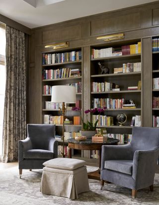Seating area in neutral shades with two upholstered armchairs, side table, floor lamp and footstool with fitted shelving behind and rug on floor