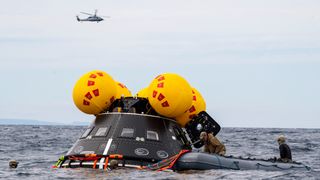 a cone-shaped spacecraft in the ocean surrounded by divers, some on a life raft. in the sky above is a large helicopter