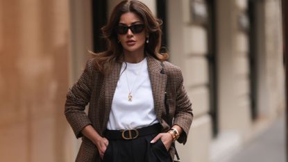 Füsun Lindner seen wearing Arket brown / black checked blazer jacket, black sunglasses, COS white cotton t-shirt, COS black wide leg pants, Ole Lynggaard gold elephant pendant long necklace and gold earrings, Hermès gold bracelets, Dior black leather belt, Louis Vuitton brown / black monogram canvas crossbody bag, during Paris Fashion Week - Womenswear Fall Winter 2023 2024, on March 03, 2023 in Paris, France. (Photo by Jeremy Moeller/Getty Images)
