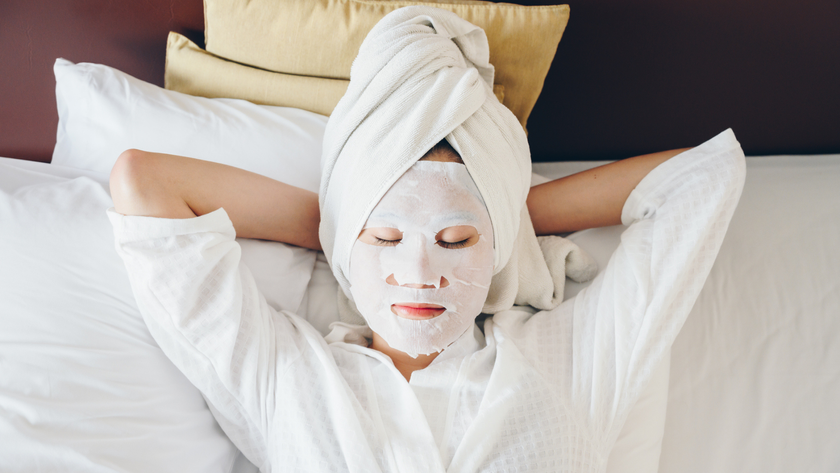 A woman sleeping on her back in bed with a sheet mask on her face and her hair wrapped in a towel