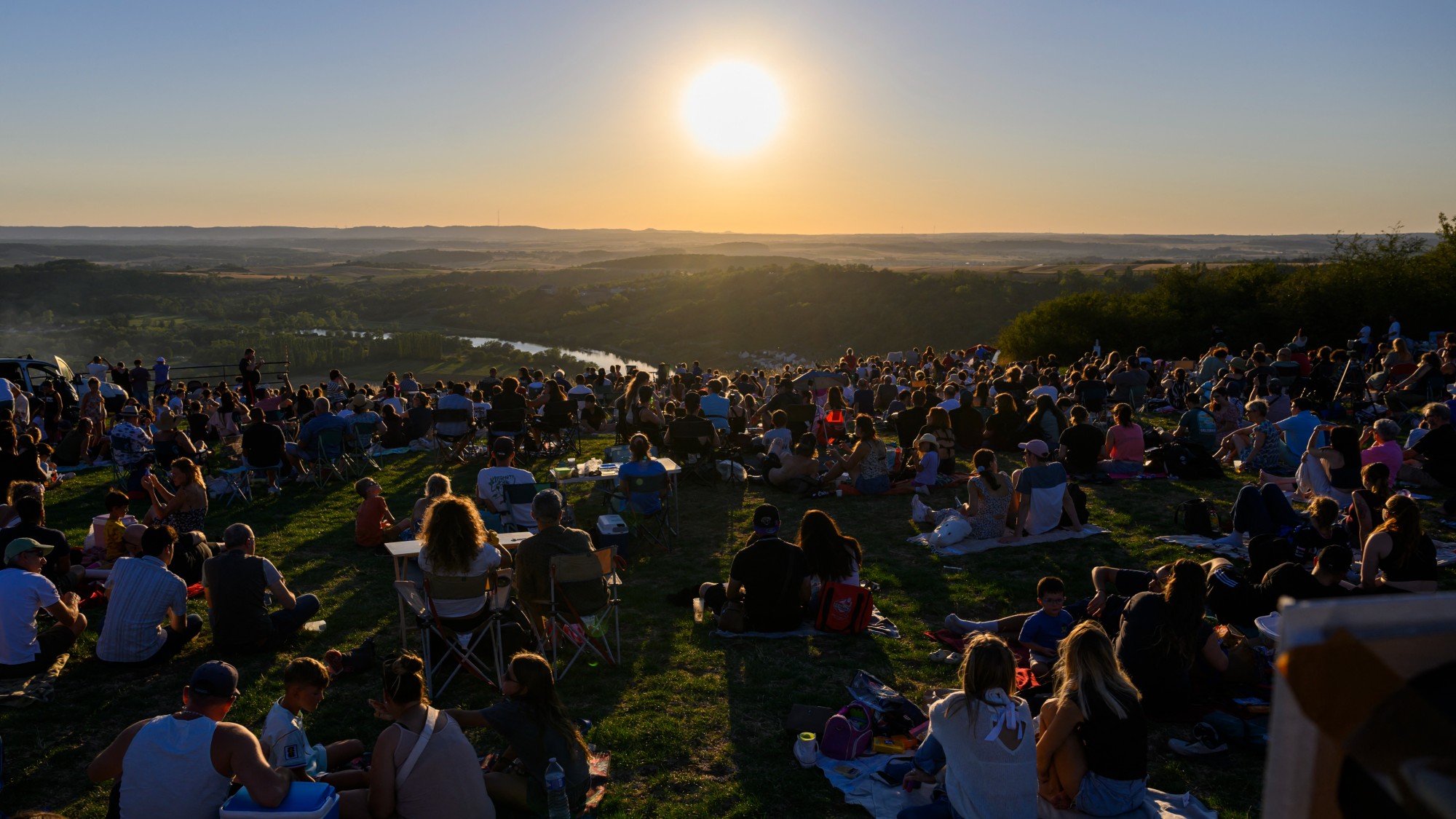 a large crowd of people sit on a hillside watching the sun set
