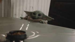 Grogu sitting in a large chair at a table and looking absolutely adorable in The Mandalorian and Grogu. 