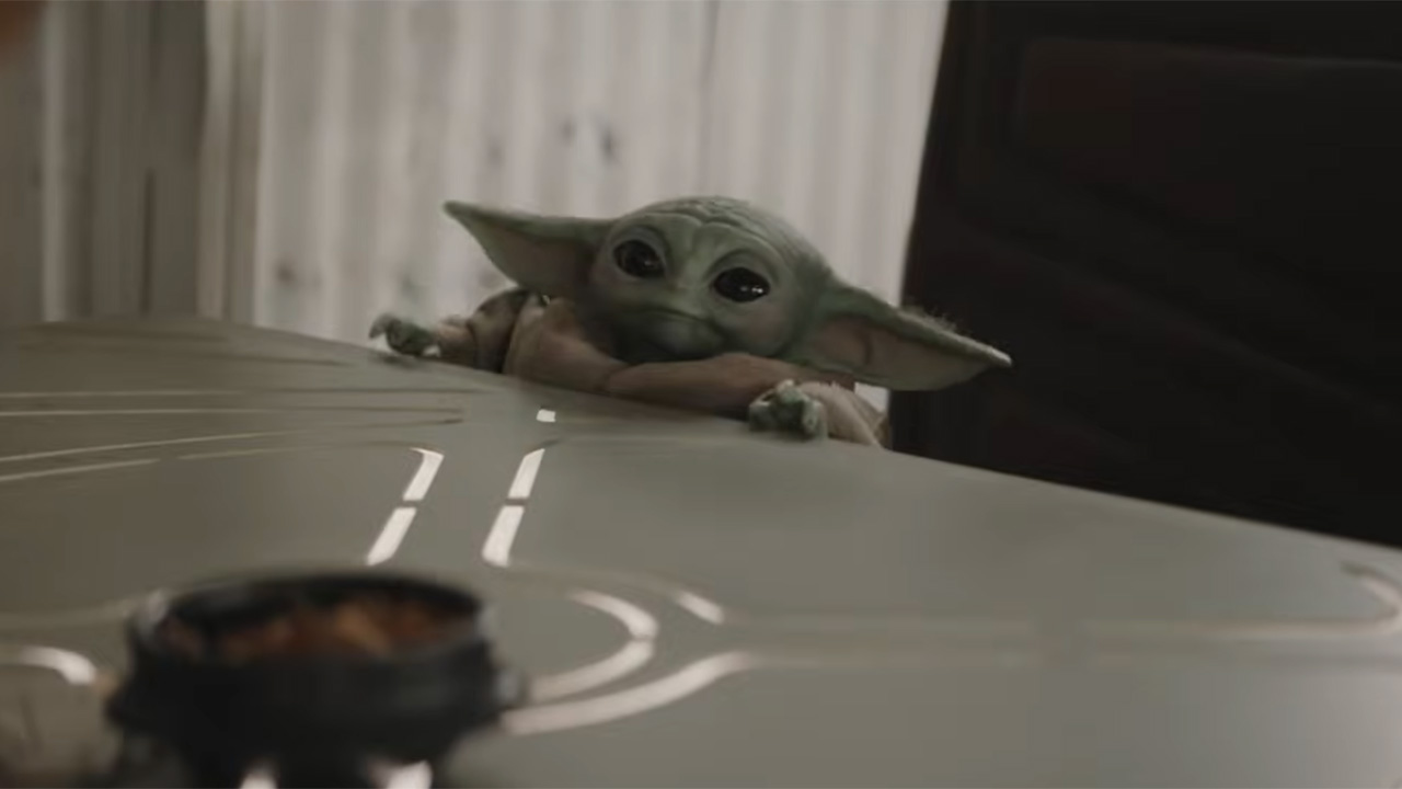 Grogu sitting in a large chair at a table and looking absolutely adorable in The Mandalorian and Grogu. 