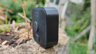 En svart Blink Outdoor-kamera står placerad på en stubbe utomhus, sedd från sidan.