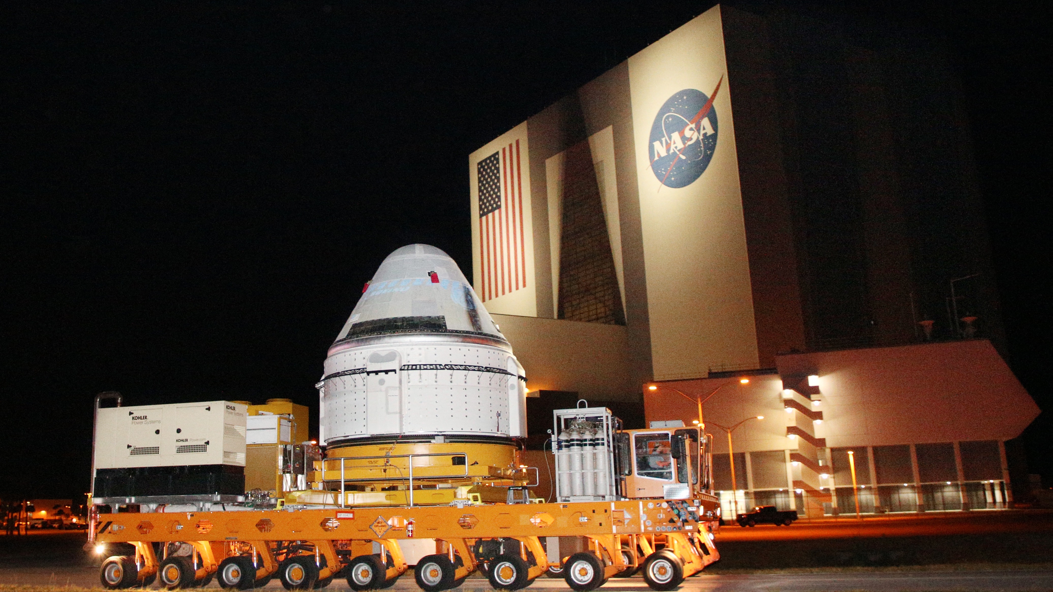 a cone-shaped spacecraft on a long trailer. in back is a large building with an american flag and nasa logo on the side
