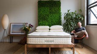 Avocado Green Mattress review: A man in a dark grey t-shirt sits next to his wooden bed, which is topped with a cream-colored Avocado mattress