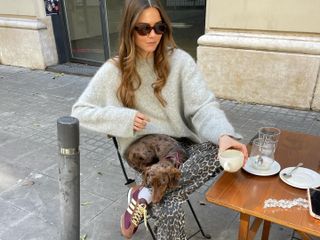 Fashion influencer @lunaisabellaa sitting outside at a cafe in Barcelona with her dog in her lap, wearing a chic spring outfit with a soft grey sweater, leopard print jeans, and Adidas SL 72 sneakers.