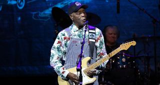 Blues icon Buddy Guy wears dungarees, a longsleeve Hawaiian shirt and ballcap as he performs with his customary Fender Stratocaster.