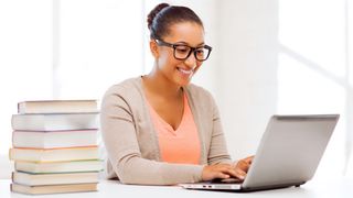 A woman wearing glasses, presumably a teacher, using a laptop at a desk next to a stack of books