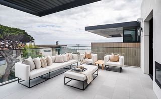 modern apartment patio with white and metal outdoor sofa and armchairs, view of the sea, pale grey floor tiles, glass panels