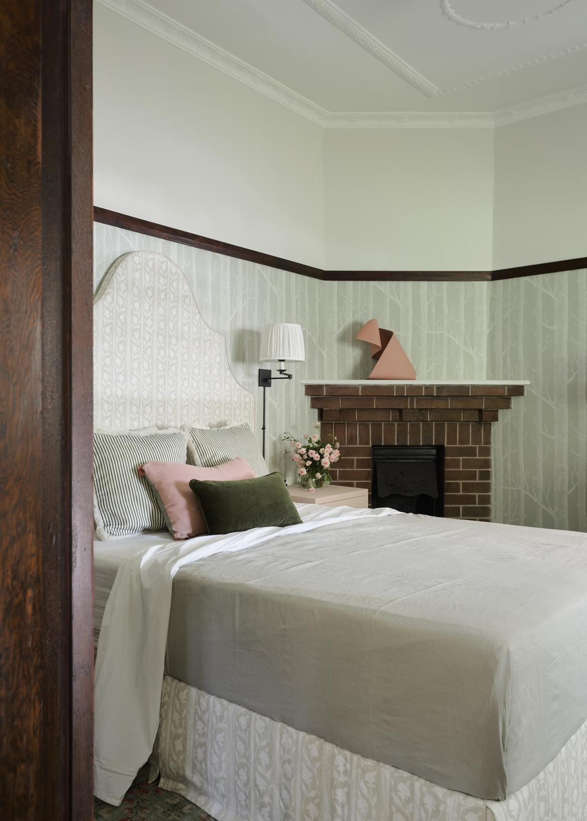 A bedroom with a brick fireplace, a traditional headboard, a patterned bed skirt, a set of colorful pillows, and an extendable wall lamp