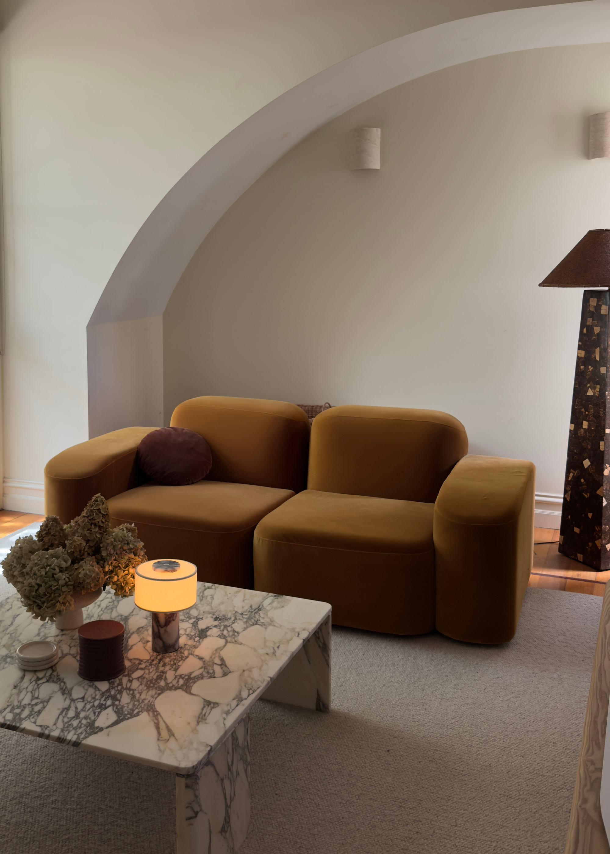 A living room with a brown sofa and large brown floor lamps with a marble styled coffee table with dried flowers, a lit portable lamp and decorative objects