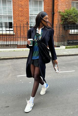 A woman wearing a navy blazer over a striped polo with a black miniskirt and white sneakers.