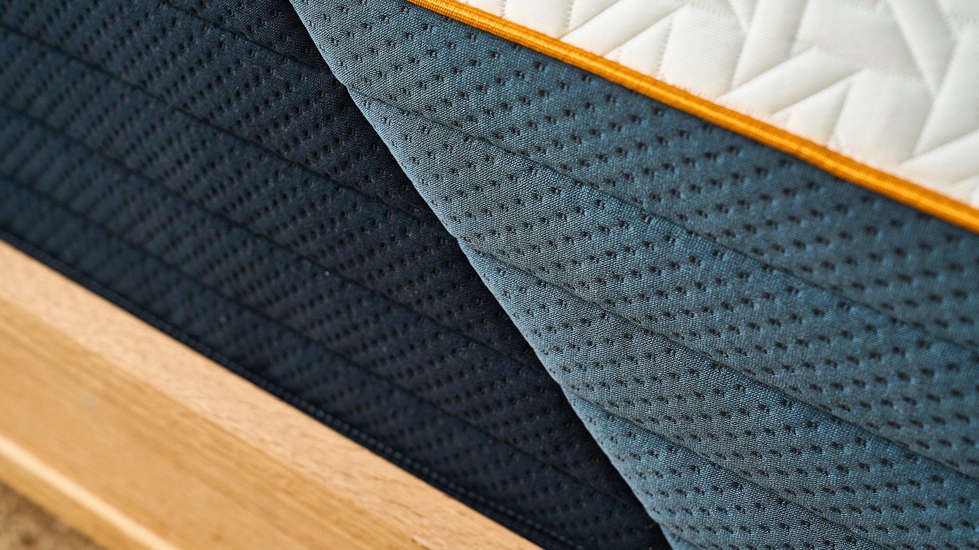 A close up detail of the fabric that makes up the soft white cover of the Nectar Ultra Hybrid mattress, the orange stitching and the light and dark blue base cover
