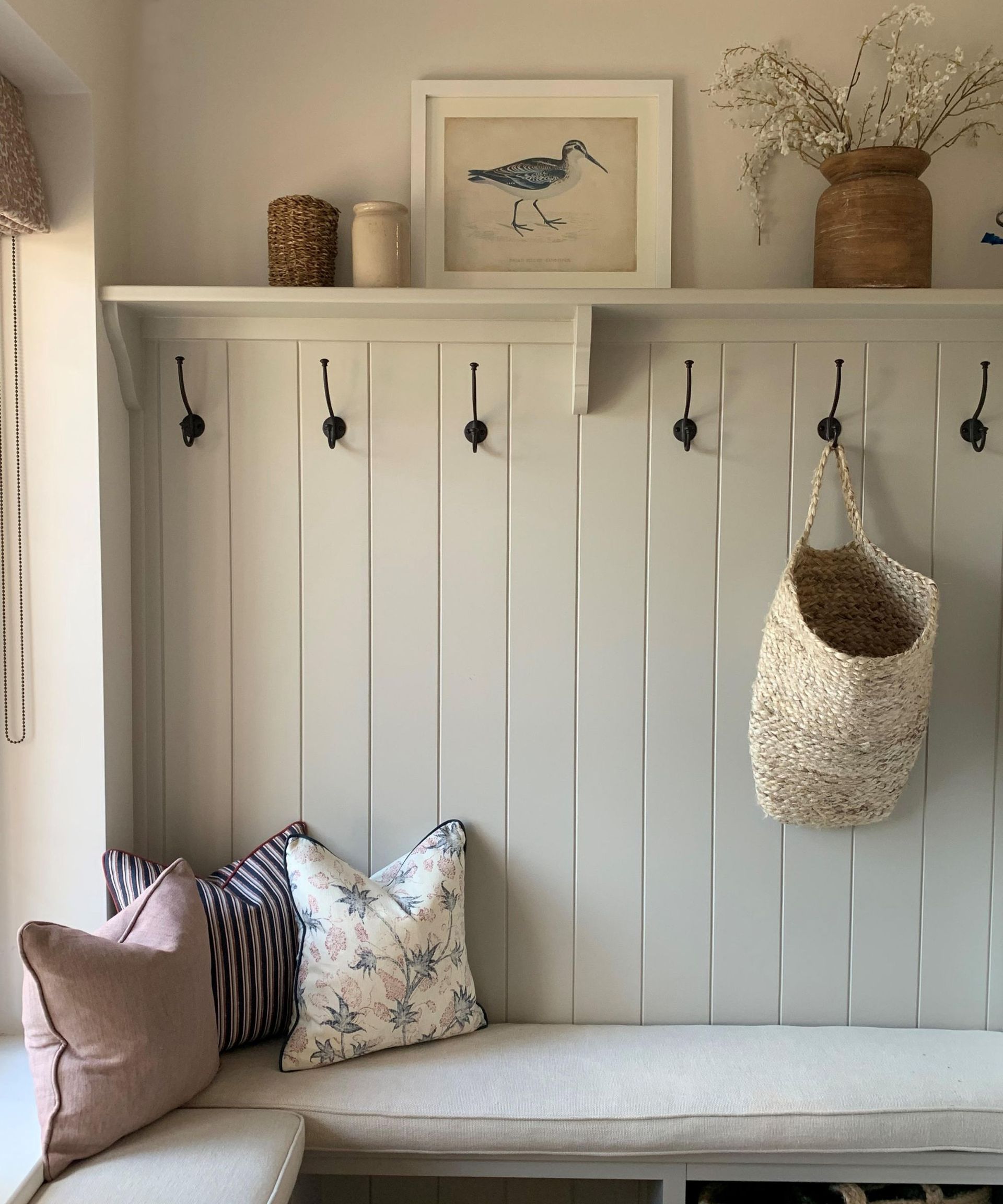 A room with white paneled walls with black hooks, a shelf with decor on it, and an L-shaped seat cushion