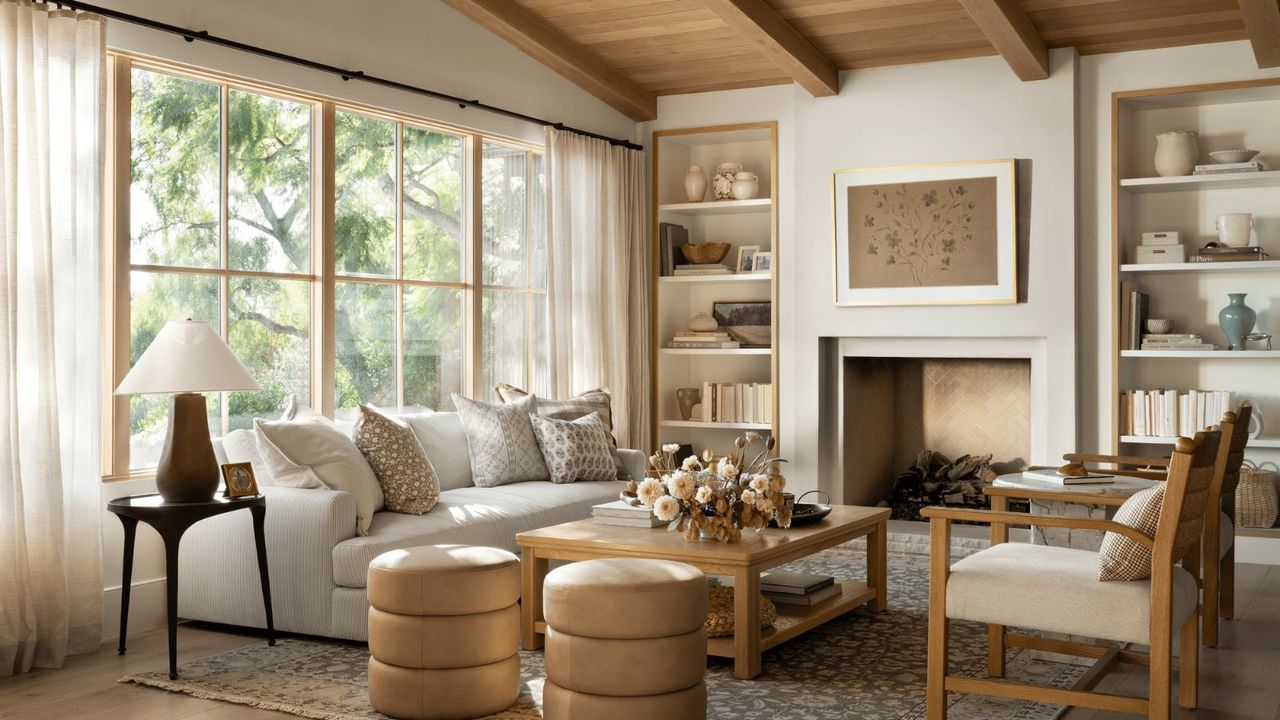 A white sofa and a wood frame love seat around a wooden coffee table, topped with a floral arrangement; in the background, a fire place set in a white wall and open shelving.