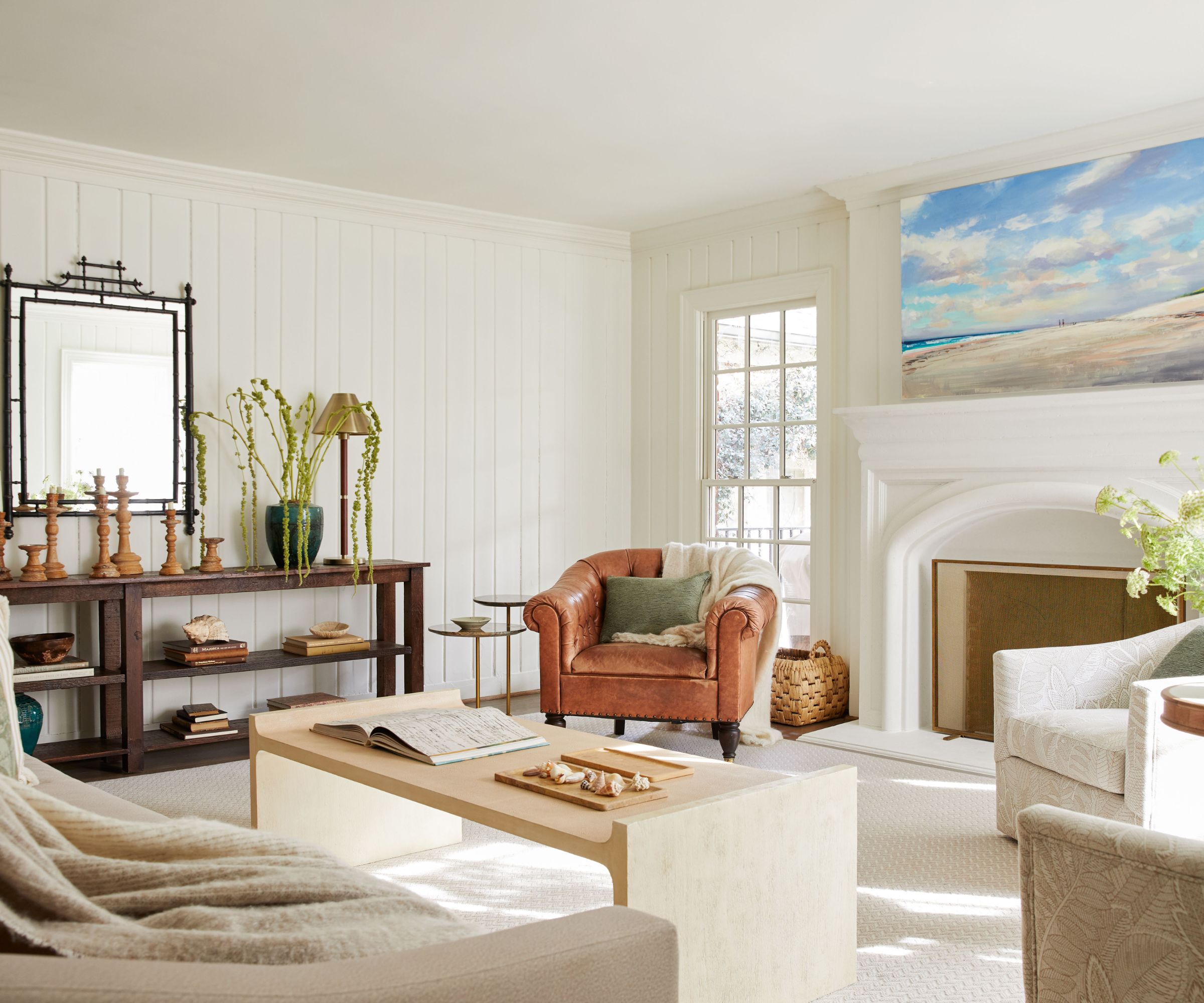 A light and airy living room with white wood-clad walls, transitional decor, a cream carpet, a white fireplace with a blue landscape artwork above it, and a tan leather armchair.