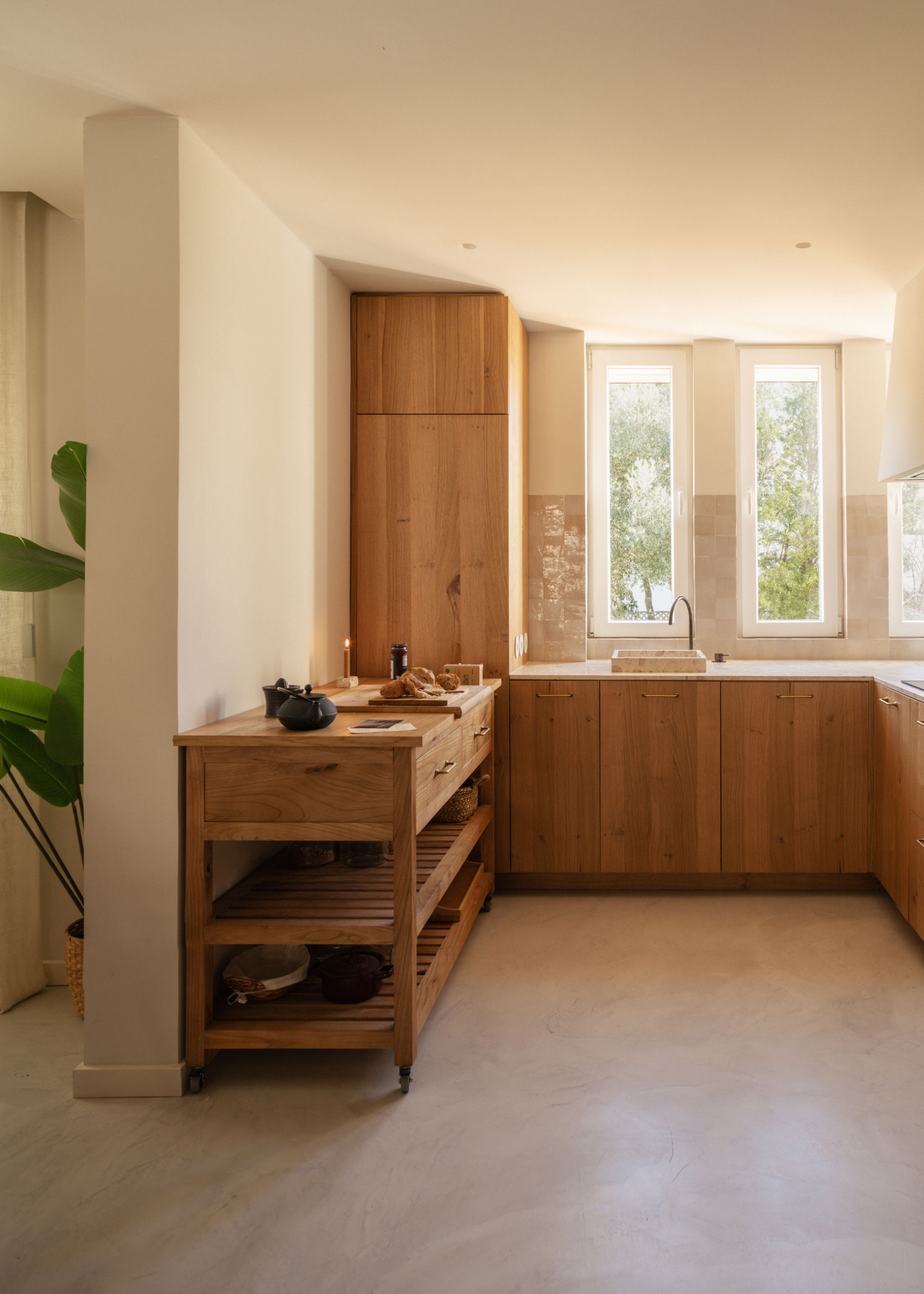 An airy white kitchen with wooden cabinets, a wooden side table with a teapot and sliced bread