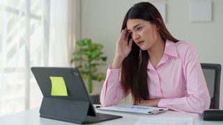 A woman sitting at a table and using a 2-in-1 laptop, holding her hand to her temple