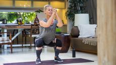 A woman in leggings, sneakers and a t-shirt practices a squat, with her hands clasped in front of her. She is exercising on a mat and behind her we see a long wooden table with chairs, couch and plant.