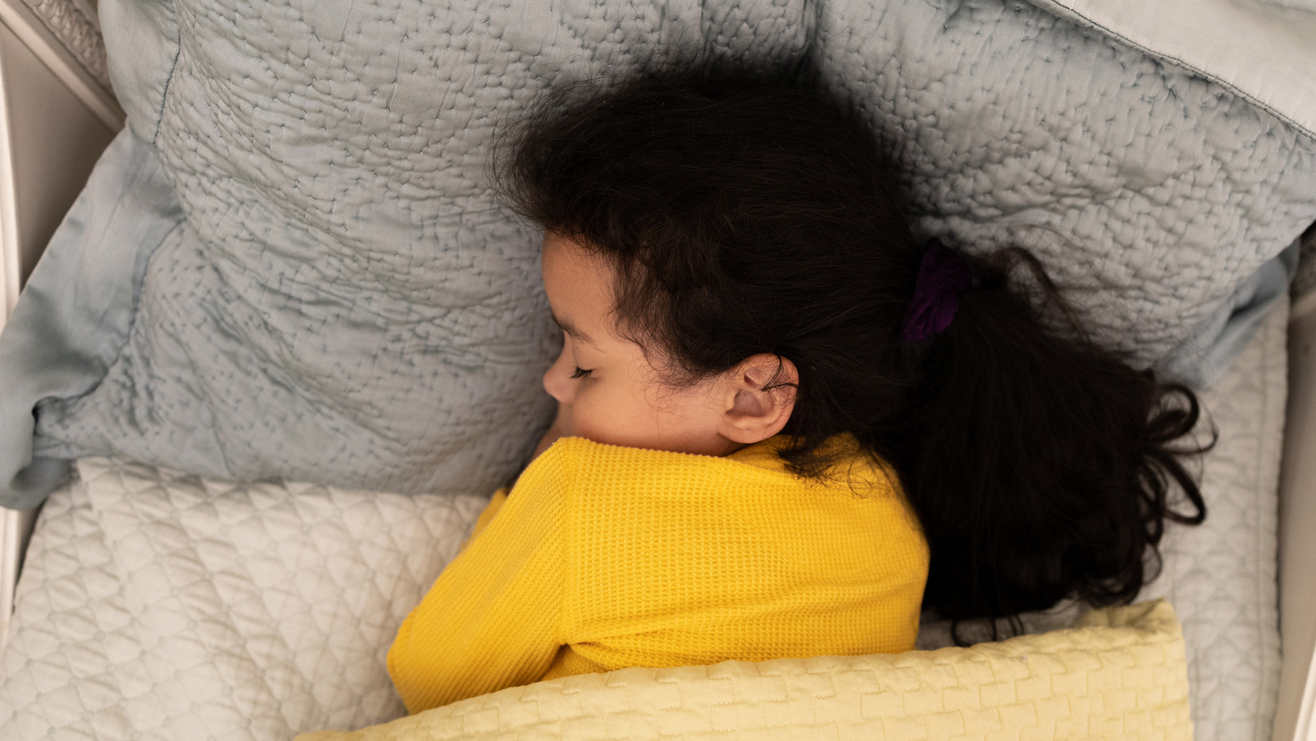A girl with long dark curly hair and a yellow shirt sleeps in a bed.