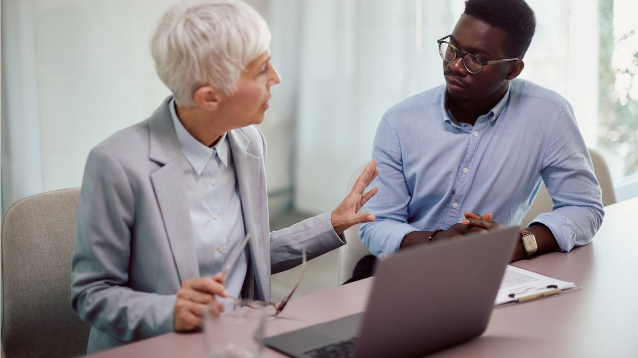 Two business people sit next to each other at a conference table, the older woman appearing to counsel the younger man.