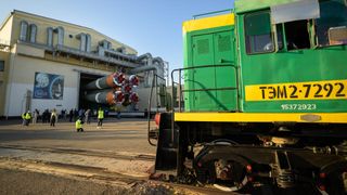 The nose of a green train car sits on the track in the foreground as gathered people in the distance - some wearing yellow, reflective vests - watch as the five-booster cluster of the butt-end of a green/grey soyuz rocket is backed out of a factory/warehouse style building. 
