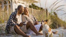 A smiling older couple sit on the beach with coffee and their dog in front of a beach house.