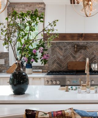 A white kitchen island with a black vase filled with green flowers and stems, a sink with gold taps, with a silver oven with gray herringbone splashback tiles behind it and white cabinets next to it and above it