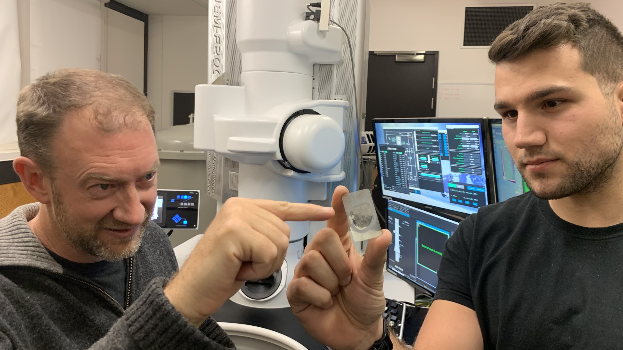 photo of two men, one holding a microscope slide, with lab tech in the background